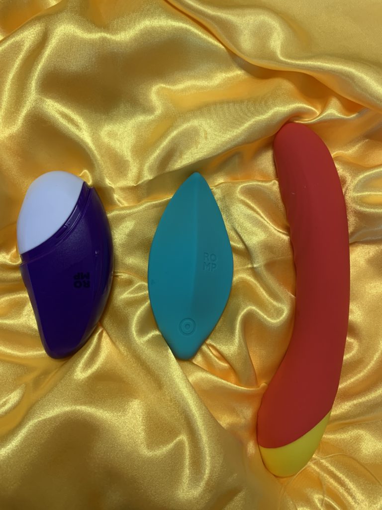 The ROMP Free, ROMP Wave, And ROMP Hype are pictured on yellow sating. The Free is an air pulse toy (oral sex suction toy), the Hype is a g-spot vibrator, and the Wave is a wearable vibe. 