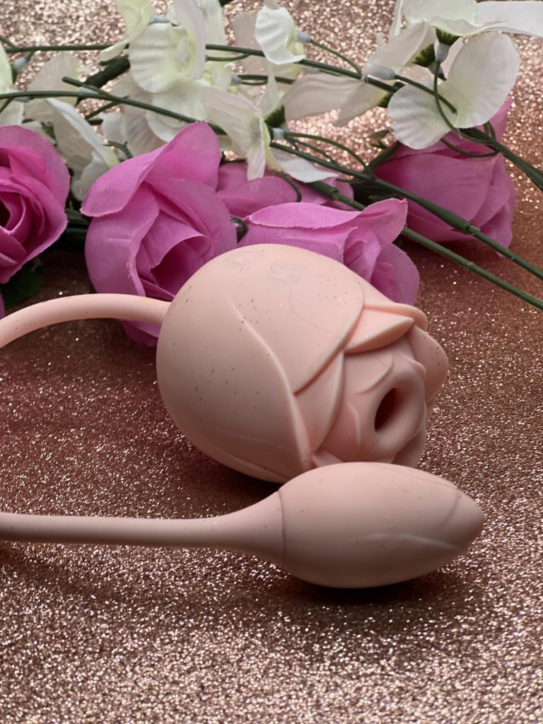 The YoYoLemon Clitoral sucker vibe is a pale pink silicone toy. It's shaped like a rose with a mouth that sucks on your clit or your nipples. In this picture, the Rose sits on a glitter background with flowers behind it
